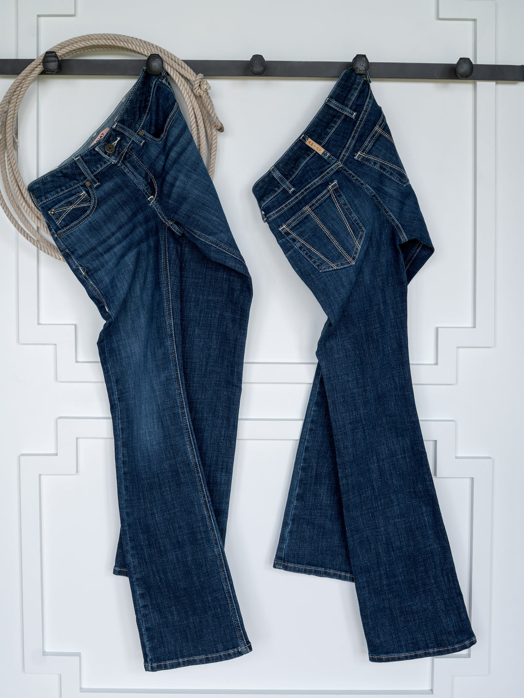 Two pairs of The Avery Perfect Rise Boot Cut Jeans by Savannah Sevens western life{&}style hang upside down by their belt loops on hooks mounted to a white wall with decorative trim. A coiled rope hangs from the leftmost hook, adding a rustic touch.