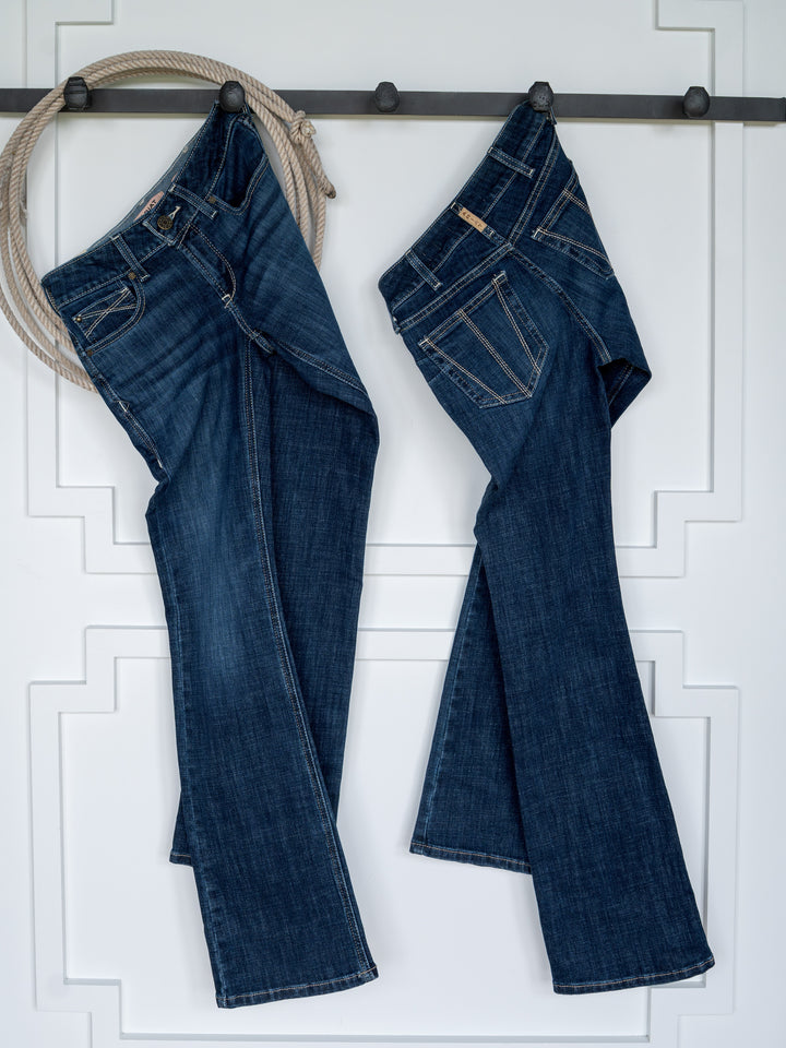 Two pairs of The Avery Perfect Rise Boot Cut Jeans by Savannah Sevens western life{&}style hang upside down by their belt loops on hooks mounted to a white wall with decorative trim. A coiled rope hangs from the leftmost hook, adding a rustic touch.