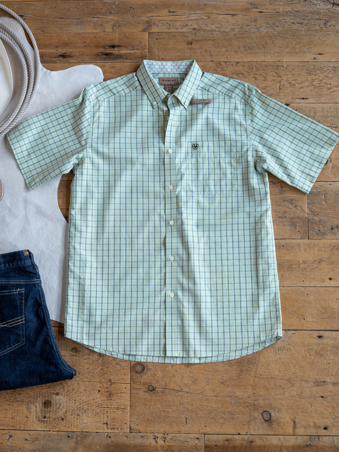 The Graham by Savannah Sevens western life{&}style is a light green men's plaid short sleeve button up shirt, shown wrinkle-free beside dark blue jeans and a white cowboy hat on a wooden floor.