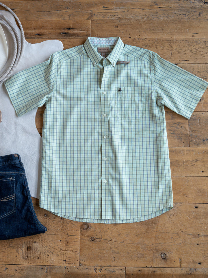 The Graham by Savannah Sevens western life{&}style is a light green men's plaid short sleeve button up shirt, shown wrinkle-free beside dark blue jeans and a white cowboy hat on a wooden floor.