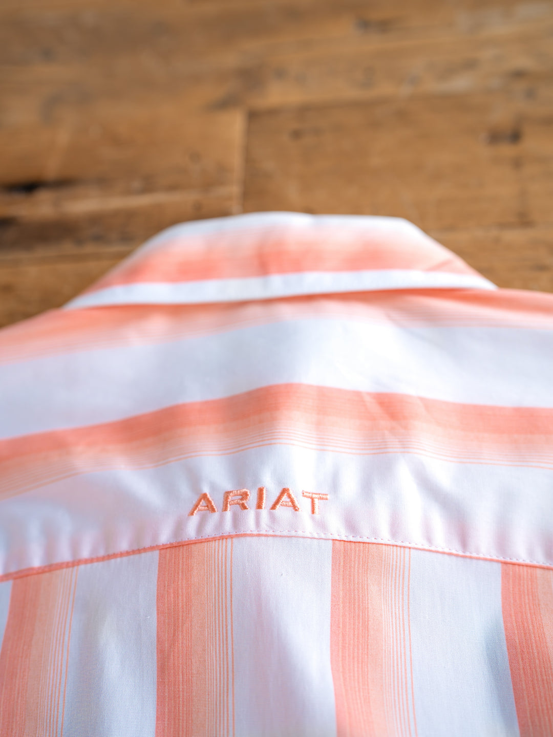A close-up of the back of The Forada, a wrinkle-resistant striped button-up by Savannah Sevens western life{&}style, with white and peach stripes and "ARIAT" embroidered in peach below the yoke, shown on a wooden surface.