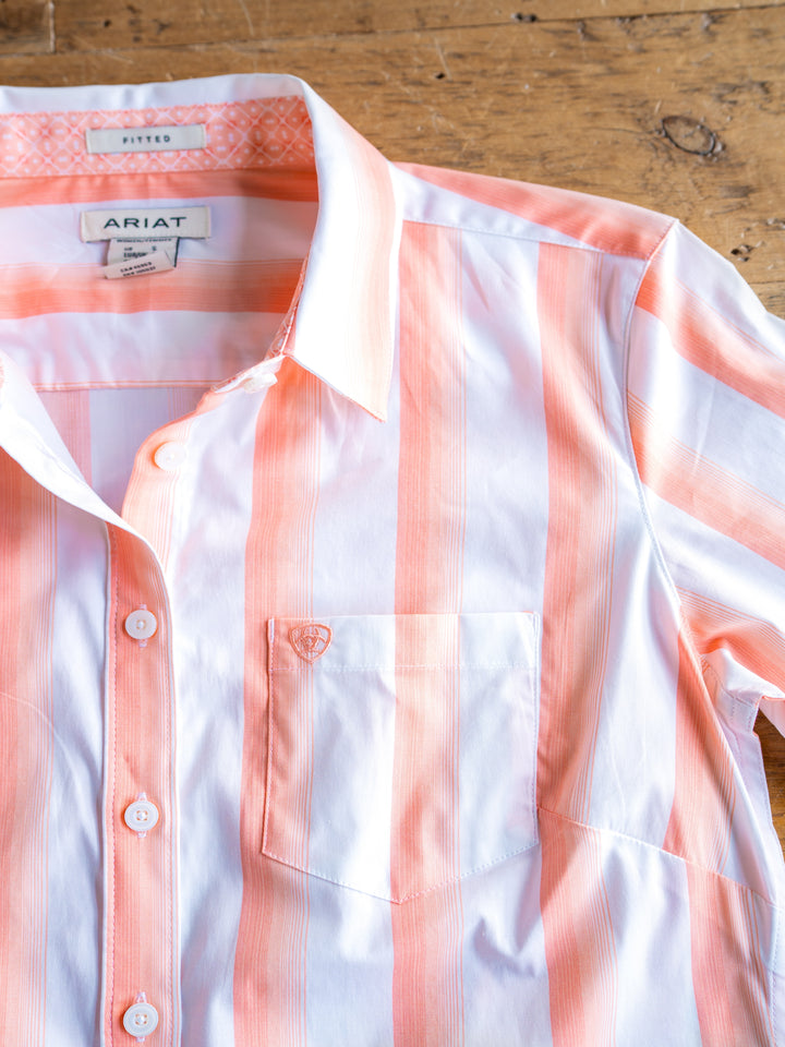 The Forada by Savannah Sevens western life{&}style is a striped button-up top with white and light orange vertical stripes, stretch fabric, collar, front pocket, and inner labels. Laid flat on a wooden surface.