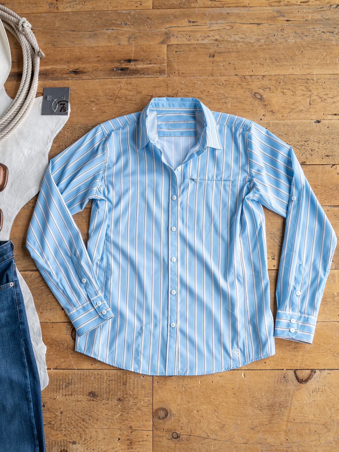 A light blue The Colmar long sleeve top by Savannah Sevens western life{&}style with white and tan stripes is laid flat on a wooden floor. Nearby are parts of a white cloth, rope, and blue jeans. Shirt features moisture wicking for comfort.