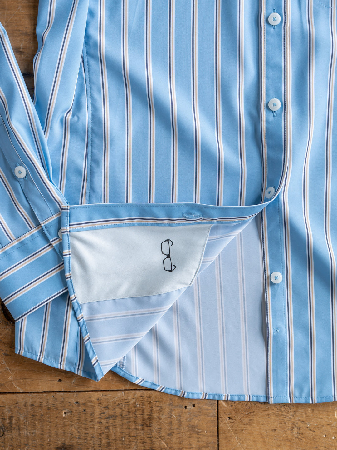 A light blue striped The Colmar long sleeve top from Savannah Sevens western life{&}style lies open on a wooden surface, showing the inside hem with a small black embroidered eyeglasses design.