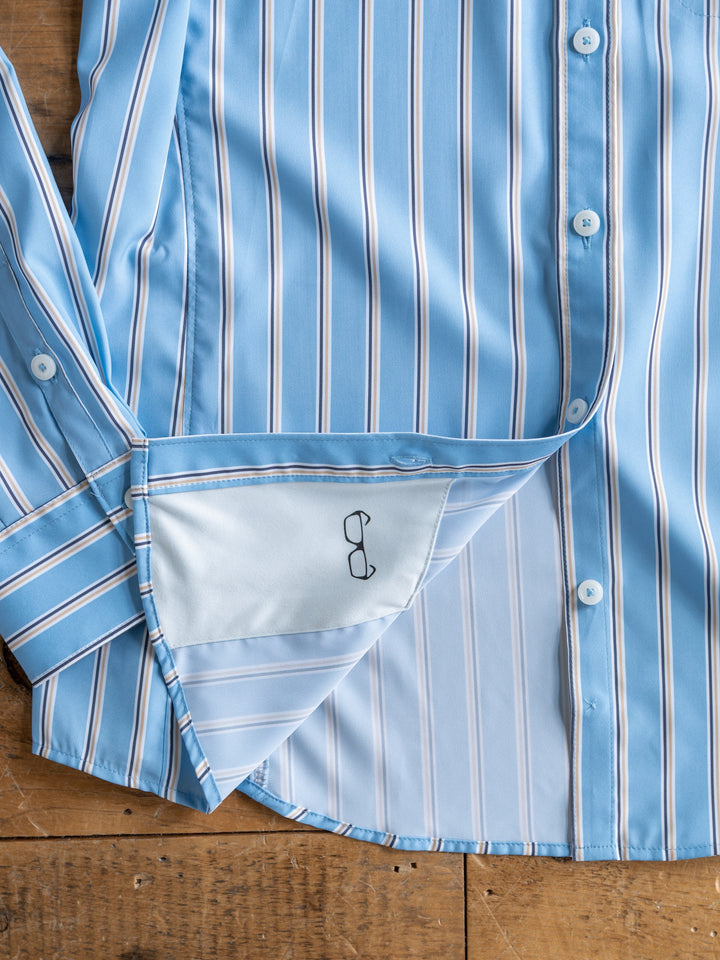 A light blue striped The Colmar long sleeve top from Savannah Sevens western life{&}style lies open on a wooden surface, showing the inside hem with a small black embroidered eyeglasses design.