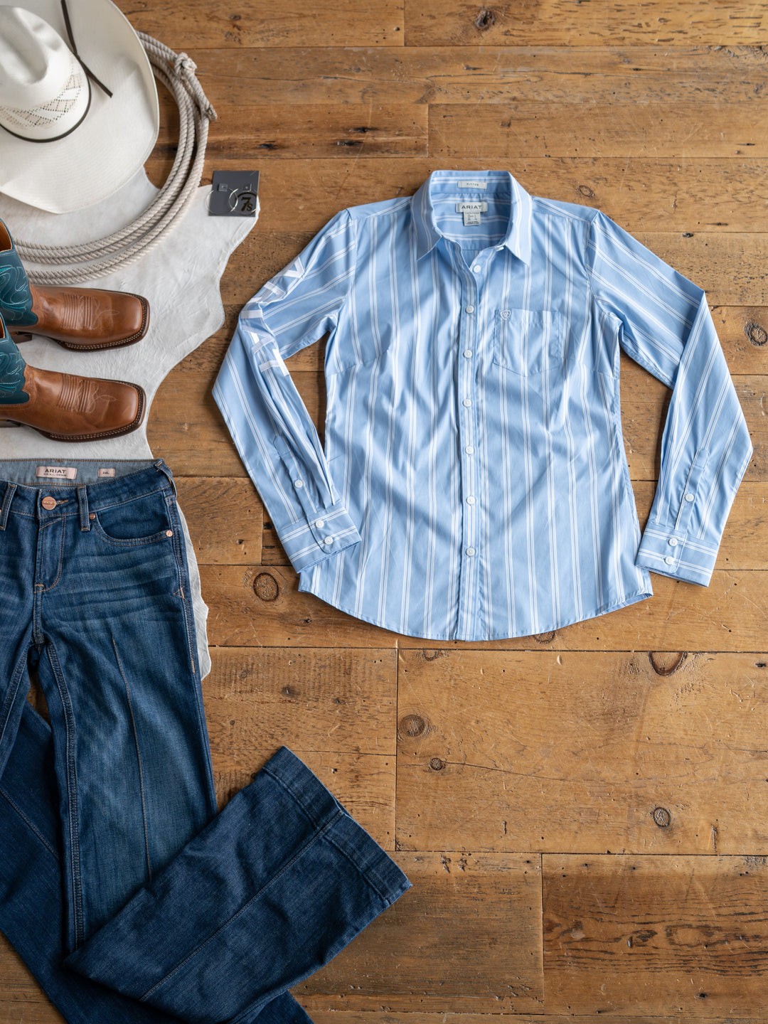 Flat lay on wood floor featuring Savannah Sevens western life{&}style's The Ingall {S-XXL}—a fitted, blue and white striped, wrinkle-resistant button up—styled with blue jeans, brown cowboy boots, a white cowboy hat, and a white scarf.