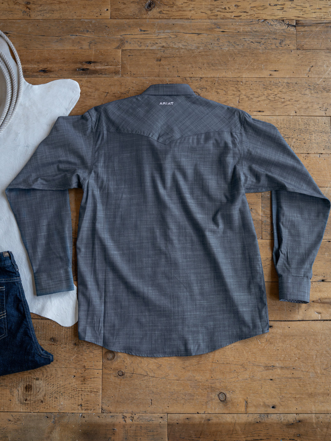 The Elkport men's shirt by Savannah Sevens western life&style is shown laid flat, back view, on a wooden floor with a white cowhide rug, striped hat, and dark denim jeans partially visible nearby.