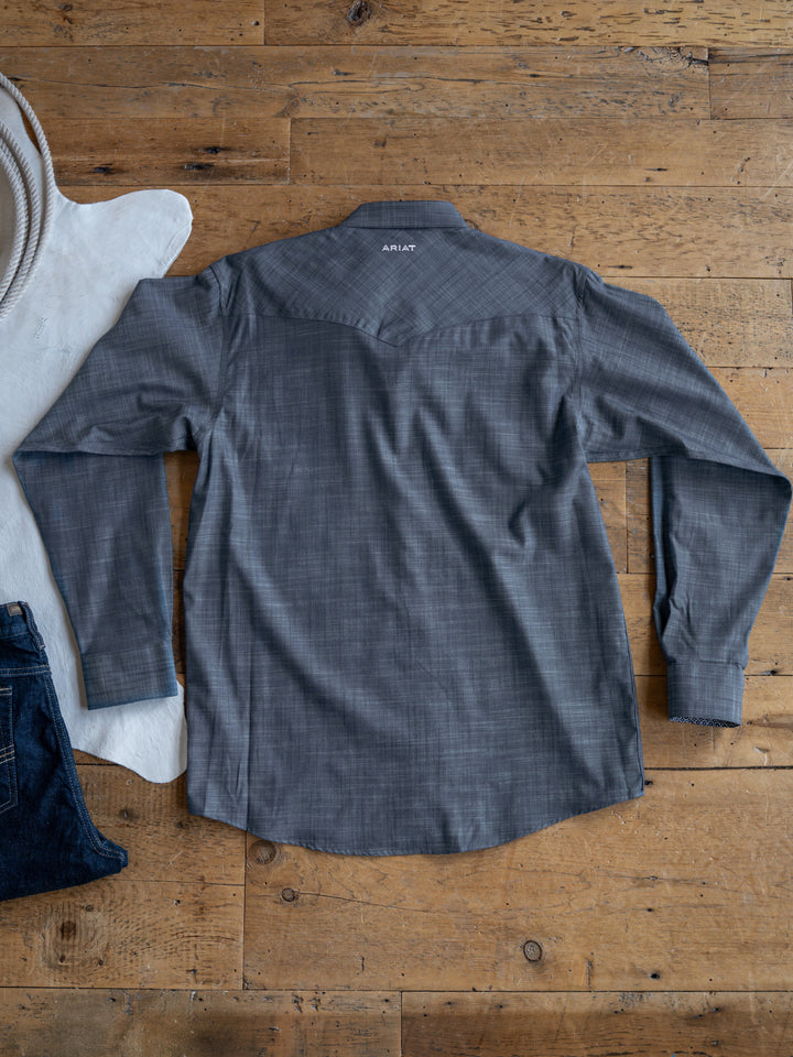 The Elkport men's shirt by Savannah Sevens western life&style is shown laid flat, back view, on a wooden floor with a white cowhide rug, striped hat, and dark denim jeans partially visible nearby.