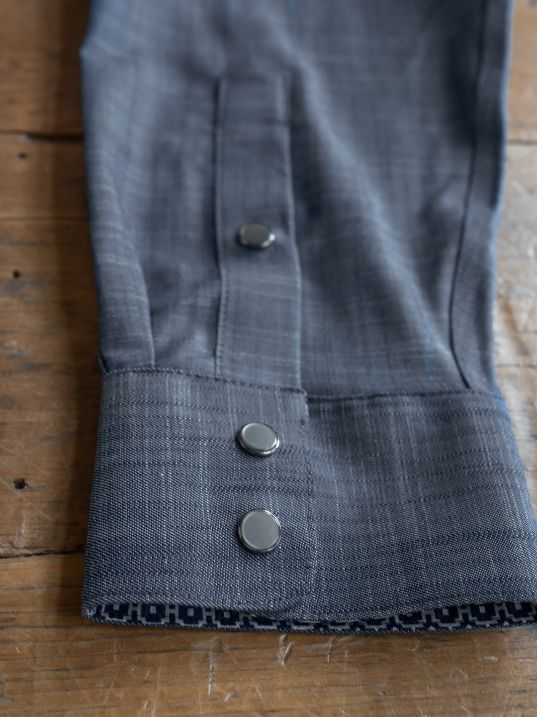 A close-up of The Elkport Men's gray suit sleeve by Savannah Sevens western life&style shows three metallic cuff buttons and a blue patterned lining—ideal with a wrinkle-free cotton shirt on a wooden surface.