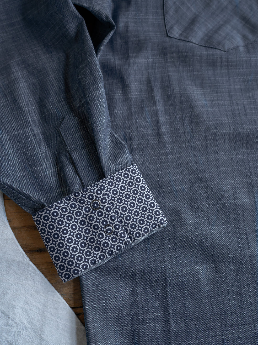 A close-up of The Elkport Men's shirt by Savannah Sevens western life&style, in blue-gray with snap buttons and a geometric inner cuff for contrast, laid flat on a wooden surface.