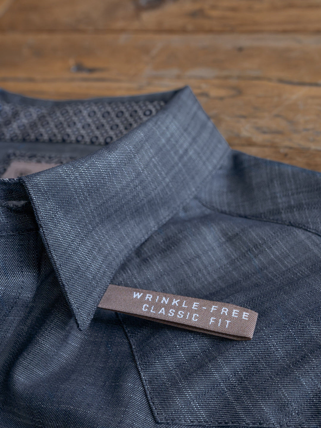 A close-up of The Elkport Men’s wrinkle-free cotton shirt by Savannah Sevens western life&style, featuring a brown label reading WRINKLE FREE CLASSIC FIT, displayed on a wooden surface.