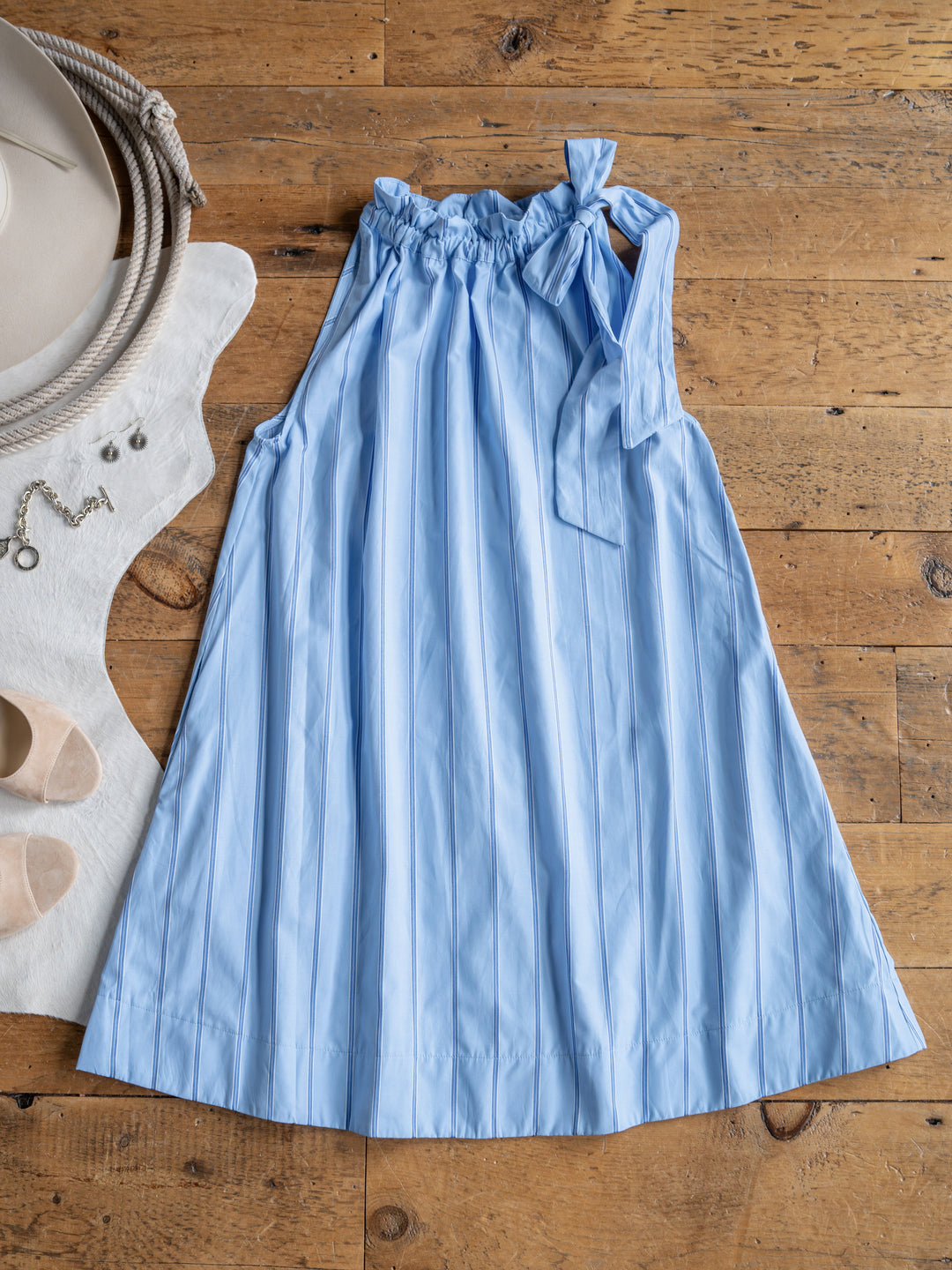 The Berkeley by Savannah Sevens western life{&}style is a light blue, sleeveless cotton blend dress with subtle vertical stripes, a ruffled neckline, and a bow on one shoulder, styled with a beige hat, jewelry, and light shoes on a wooden floor.