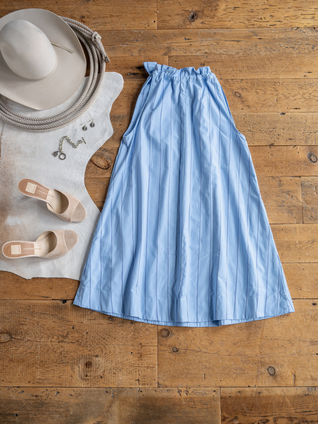 The Berkeley dress by Savannah Sevens western life{&}style is displayed on a wooden floor beside beige heels, a wide-brimmed hat, necklace, and earrings, all styled atop a white fur rug with a coiled rope.