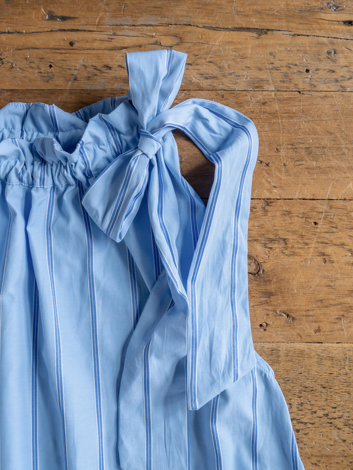 The Berkeley by Savannah Sevens western life{&}style: a light blue cotton-blend dress with white and blue vertical stripes and a large bow on one shoulder, shown flat on a rustic wooden surface.