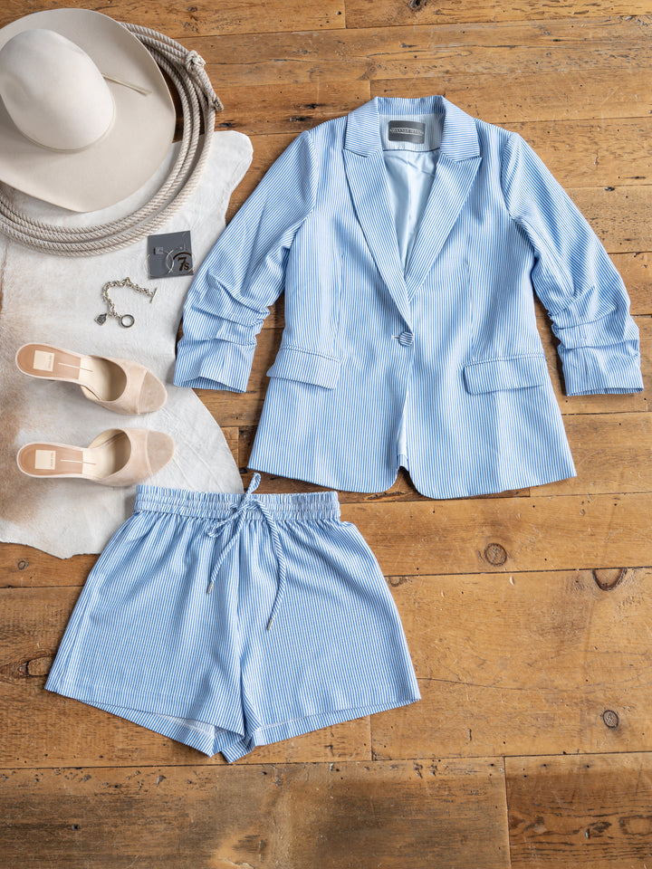 A flat lay on a wooden floor features The Malin {Set} by Savannah Sevens western life{&}style—blue and white pinstripe blazer with matching high waisted shorts—styled with beige heels, a wide-brim hat, and a key charm bracelet on a cowhide rug.