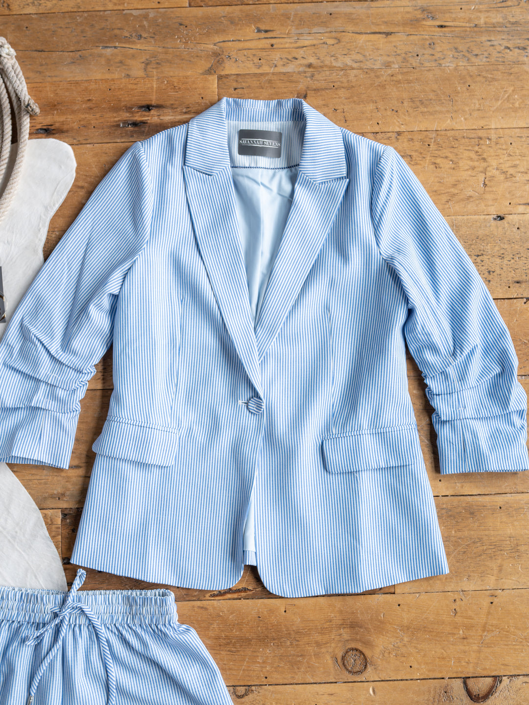 The Malin {Set} by Savannah Sevens western life{&}style, a blue and white pinstriped single-button blazer paired with matching high-waisted shorts, is displayed flat on a wooden floor beside a coiled rope.