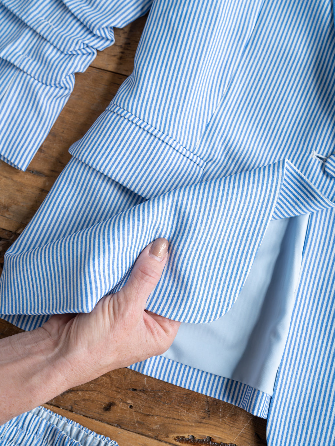 A hand lifts the edge of The Malin {Set} blue and white pinstripe blazer by Savannah Sevens western life{&}style, revealing its light blue lining on a wooden surface.