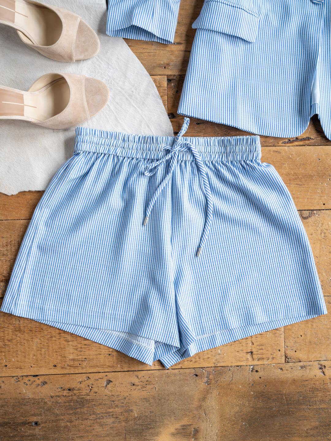 The Malin {Set} by Savannah Sevens western life{&}style features light blue and white pinstriped high-waisted pajama shorts with a drawstring waist, shown beside the matching top and beige shoes on a wooden floor.