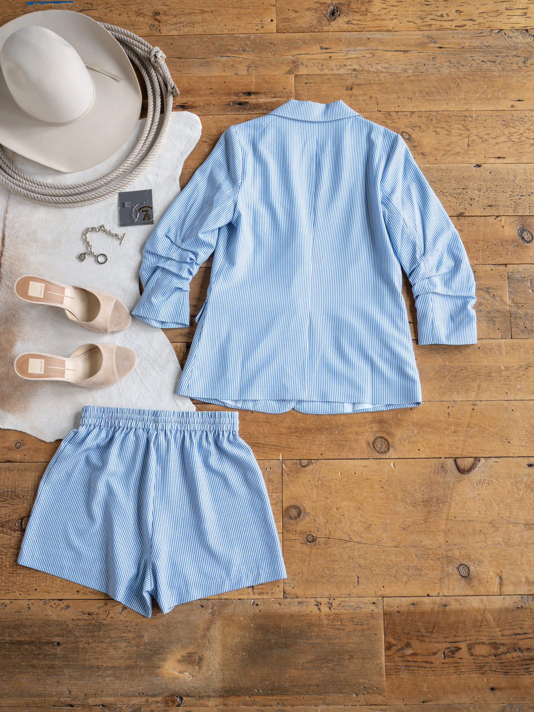 The Malin {Set} by Savannah Sevens western life{&}style—a blue and white pinstriped blazer and shorts with a button-up top—is displayed on a wooden floor with beige slip-ons, a wide-brim hat, bracelet, and earrings nearby.