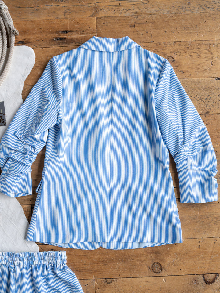 The Malin {Set} by Savannah Sevens western life{&}style features a light blue and white vertically striped long-sleeve shirt laid flat, back up, on wood, with part of the matching high-waisted shorts from the pinstriped set visible at the edge.