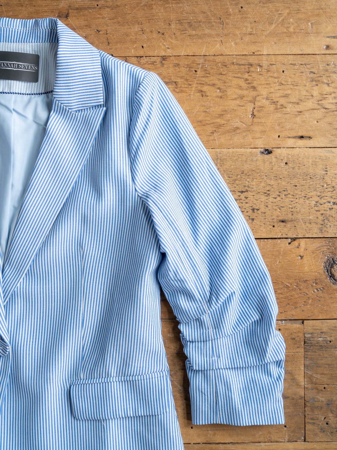 The Malin {Set} by Savannah Sevens western life{&}style features a blue and white pinstripe blazer with ruched sleeves, shown on a wooden surface with only its left half visible.