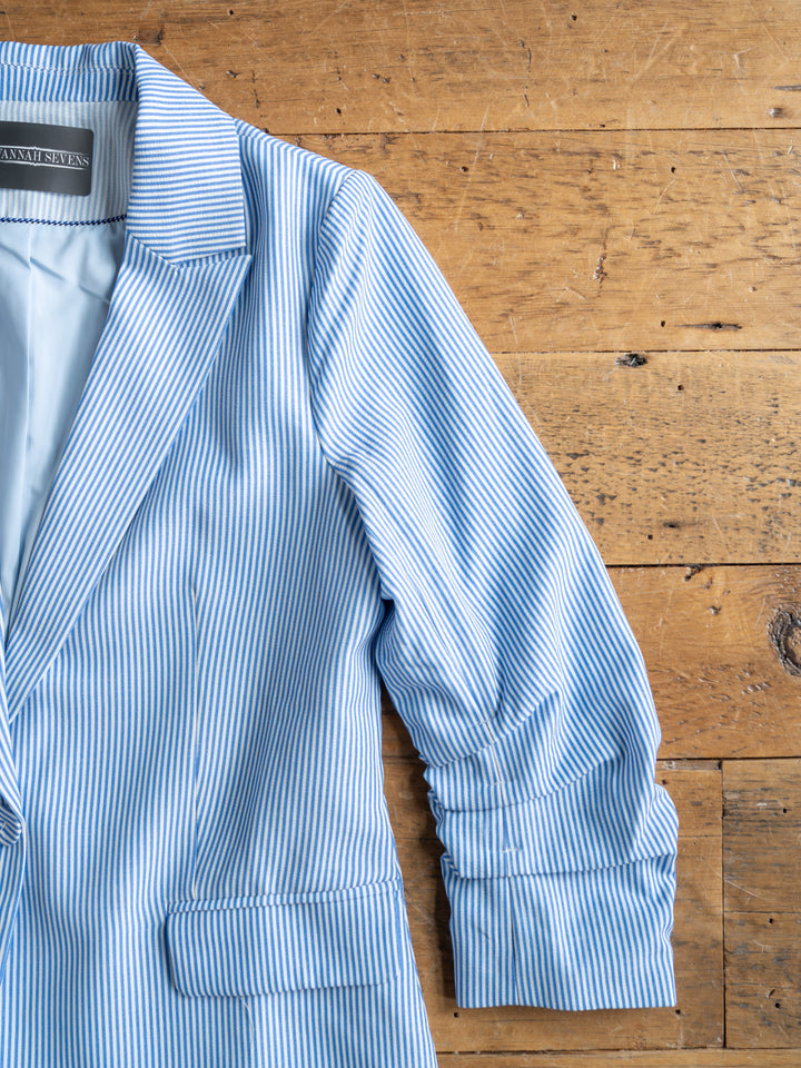 The Malin {Set} by Savannah Sevens western life{&}style features a blue and white pinstripe blazer with ruched sleeves, shown on a wooden surface with only its left half visible.