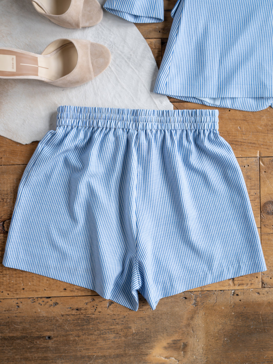 The Malin {Set} from Savannah Sevens western life{&}style features light blue and white pinstriped high-waisted shorts with an elastic waistband, laid flat on wood, with beige heels and a matching blazer partially visible nearby.
