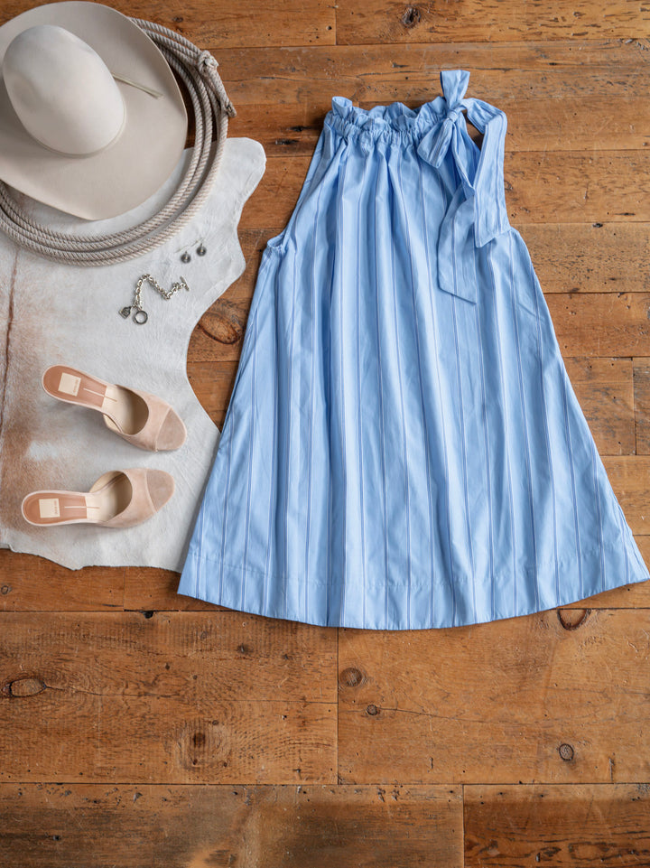 The Berkeley by Savannah Sevens western life{&}style—a light blue cotton blend dress—is styled on a wooden floor with a beige wide-brimmed hat, silver jewelry, nude heels, and a cowhide rug.