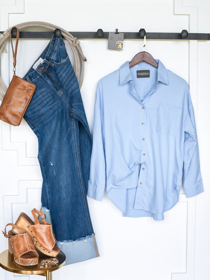 The Driscoll jeans by Savannah Sevens western life{&}style, an ice blue pinstripe button-up, tan heeled sandals, and a brown leather purse hang on a black wall rack against a white background.