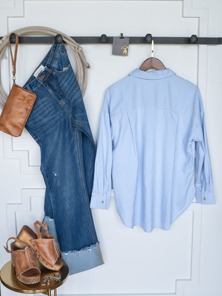 The Driscoll jeans by Savannah Sevens western life{&}style, an ice blue pinstripe button-up, tan heeled sandals, and a brown leather crossbody bag hang on hooks above a gold table against a white wall.