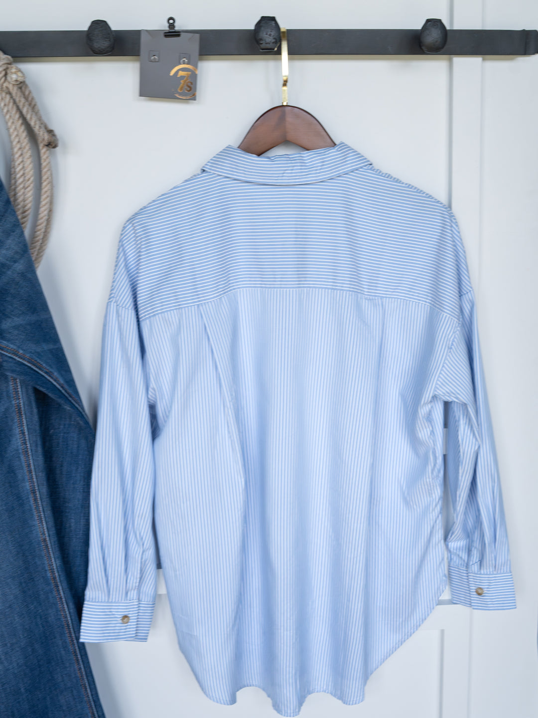 The Driscoll by Savannah Sevens western life{&}style, an ice blue and white pinstripe oversized button-up top, hangs on a wooden hanger from a black coat rack. A denim jacket is partially visible to the left, with a rope on the same rack.