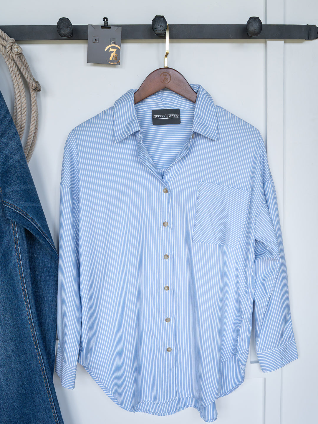 The Driscoll by Savannah Sevens western life{&}style, an oversized relaxed fit ice blue and white pinstripe button-up top, hangs on a wooden hanger from a black metal rack beside a denim jacket and thick rope.
