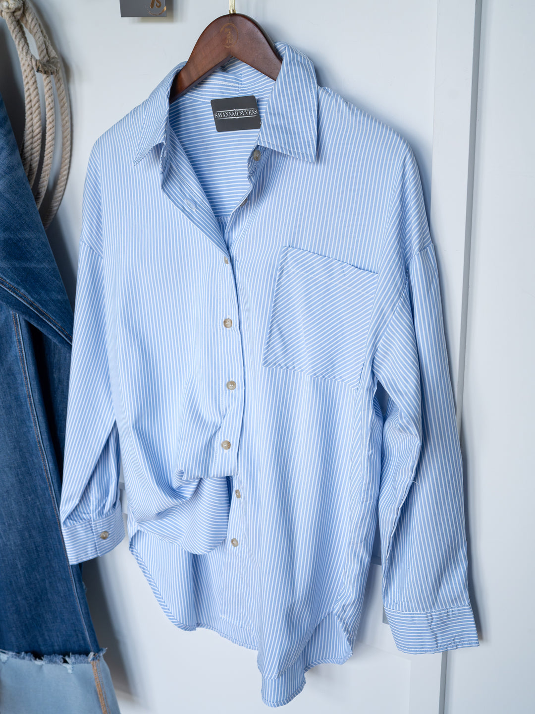 The Driscoll by Savannah Sevens western life{&}style is an ice blue pinstripe button up top with a relaxed, oversized fit, single chest pocket, and denim fabric, shown on a wooden hanger against a white wall.