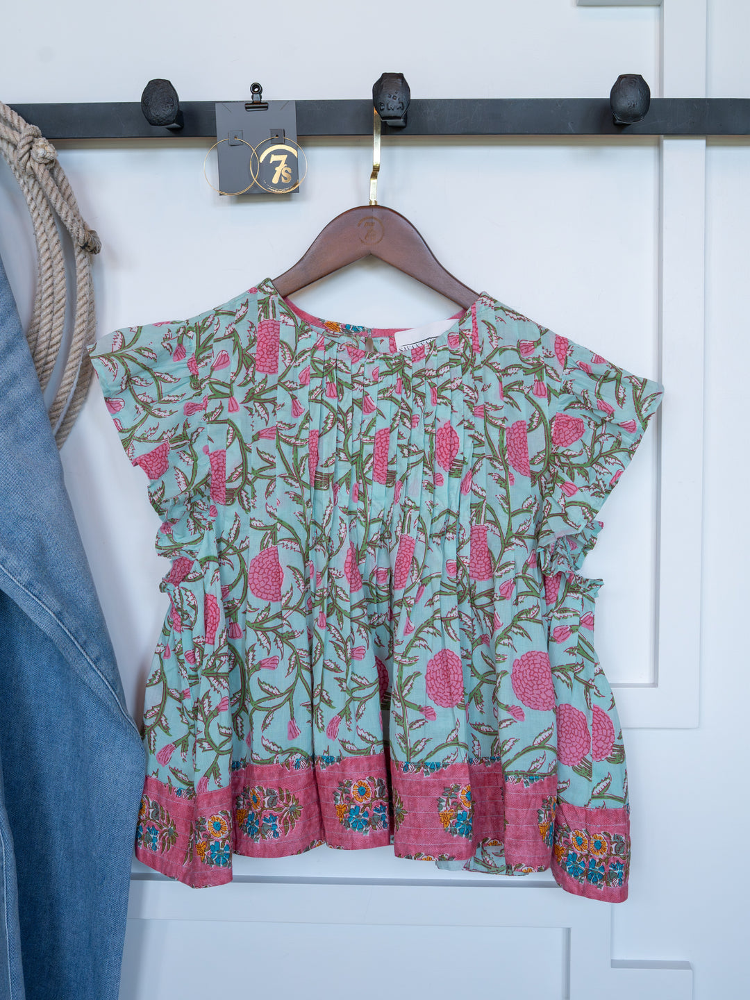 The Hartley by Savannah Sevens western life{&}style, a vibrant cotton floral top with ruffled sleeves, hangs on a wooden hanger from a black wall hook. A denim piece and rope are partially seen on the left.