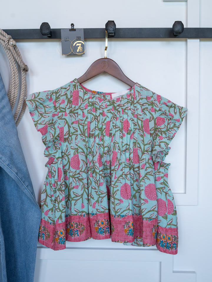 The Hartley by Savannah Sevens western life{&}style, a vibrant cotton floral top with ruffled sleeves, hangs on a wooden hanger from a black wall hook. A denim piece and rope are partially seen on the left.
