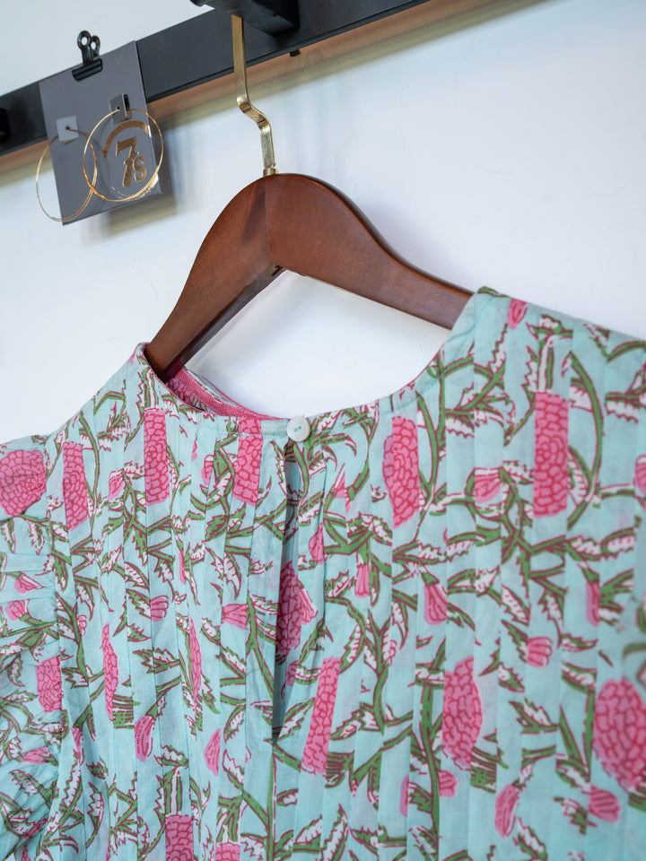 A close-up of The Hartley by Savannah Sevens western life{&}style, a cotton floral top with a pink print on light blue, hanging on a wooden hanger. It features a buttoned keyhole back and is displayed on a rack with a gold number tag.