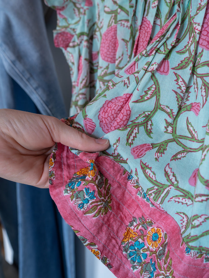 A hand holds the edge of The Hartley, a light green and pink cotton floral top with a contrasting pink border featuring blue and yellow flowers by Savannah Sevens western life{&}style. Denim fabric is seen in the background.