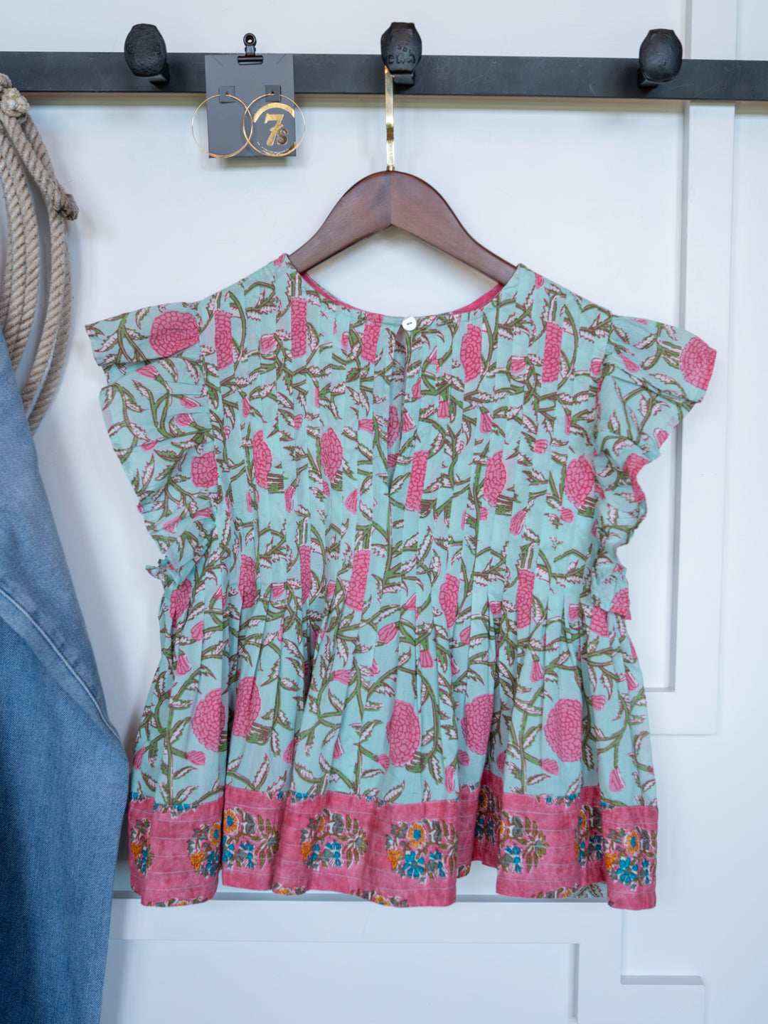 The Hartley by Savannah Sevens western life{&}style is a frilly, sleeveless cotton top for children featuring a pink and green floral design with a pink floral border, displayed on a wooden hanger against a white wall.