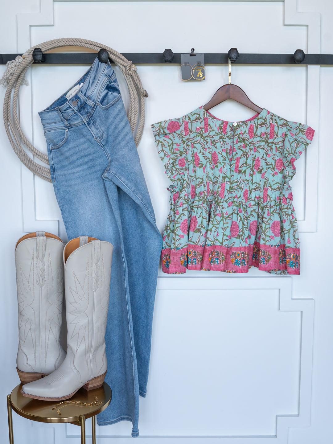 A flat lay showcases The Hartley by Savannah Sevens western life{&}style—light-wash jeans, a pink and green floral top, cream cowboy boots, and a coiled rope arranged on a white paneled wall.