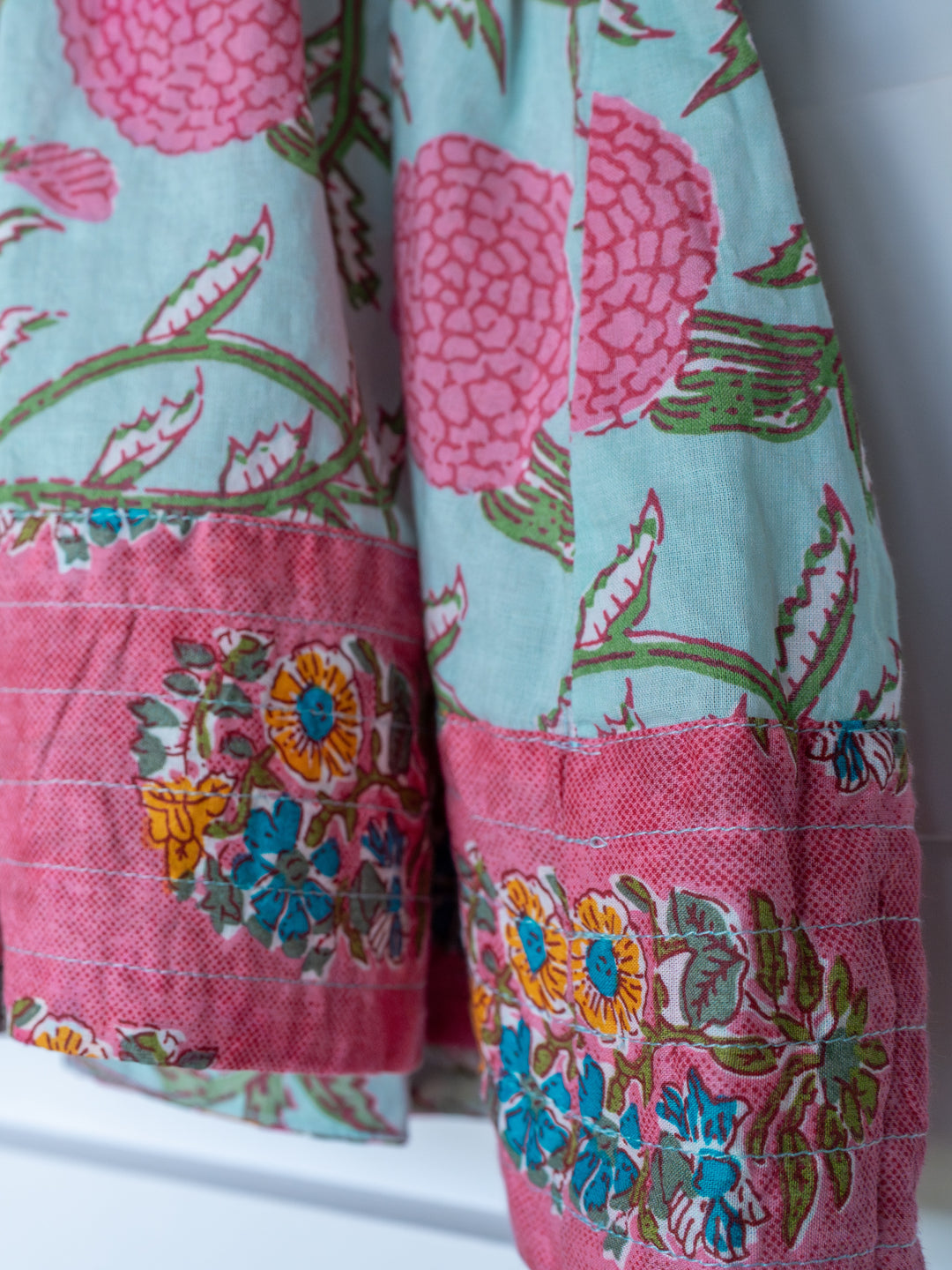 A close-up of The Hartley by Savannah Sevens western life{&}style, a cotton floral top featuring large pink flowers on light blue, finished with a contrasting pink border adorned with blue, yellow, and green flowers.