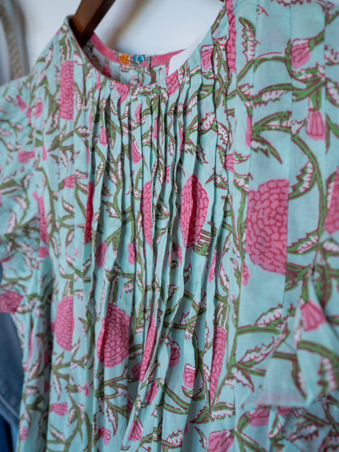 A close-up of The Hartley top by Savannah Sevens western life{&}style, a light blue cotton floral piece with pink patterns, green leaves, front pleats, and gathered neckline detail, displayed on a wooden hanger.