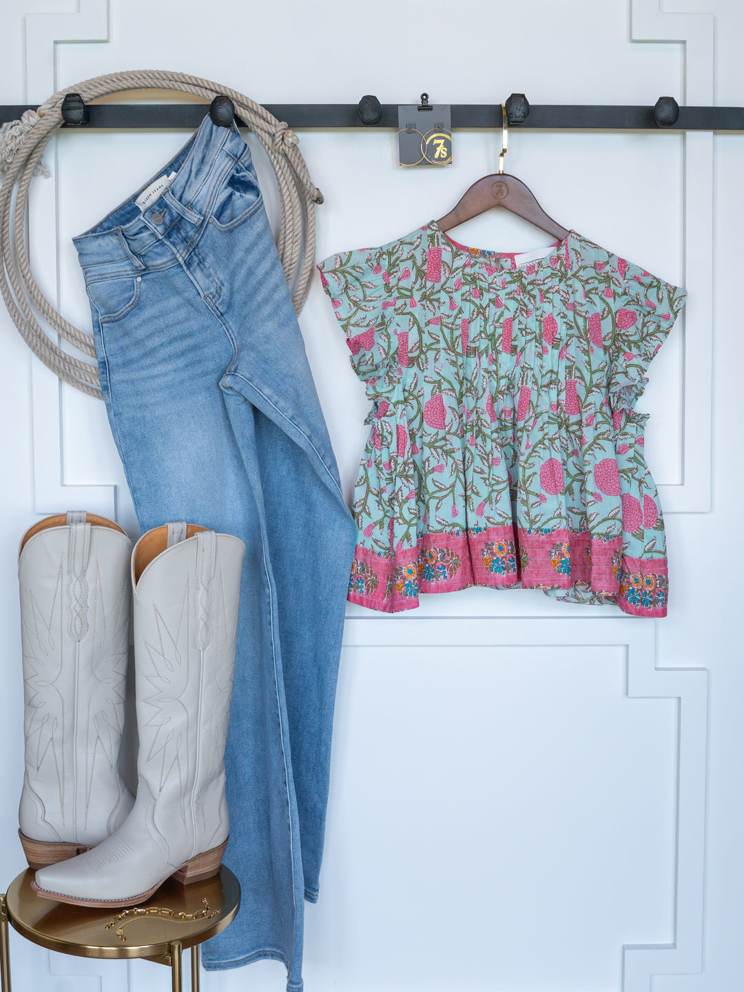 The Hartley by Savannah Sevens western life{&}style—a pair of blue jeans and a floral short sleeve top—hangs on the wall, with white cowboy boots displayed below on a small round table.