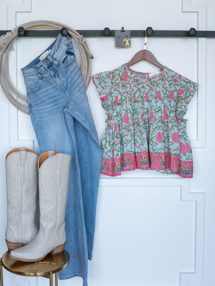 The Hartley by Savannah Sevens western life{&}style—a pair of blue jeans and a floral short sleeve top—hangs on the wall, with white cowboy boots displayed below on a small round table.