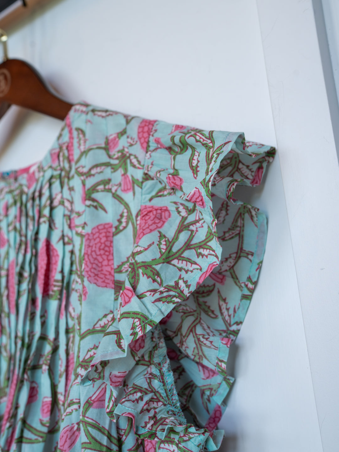 Close-up of The Hartley by Savannah Sevens western life{&}style, a light blue cotton floral top with pink and green flowers, ruffled sleeves, and pleats, displayed on a wooden hanger against a white background.