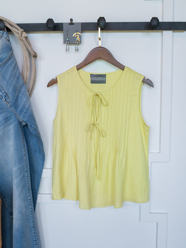 The Marlette by Savannah Sevens western life{&}style—a chartreuse pleat top with an adjustable tie neckline—hangs on a wooden hanger, with blue jeans and earrings nearby on the same black wall rack.