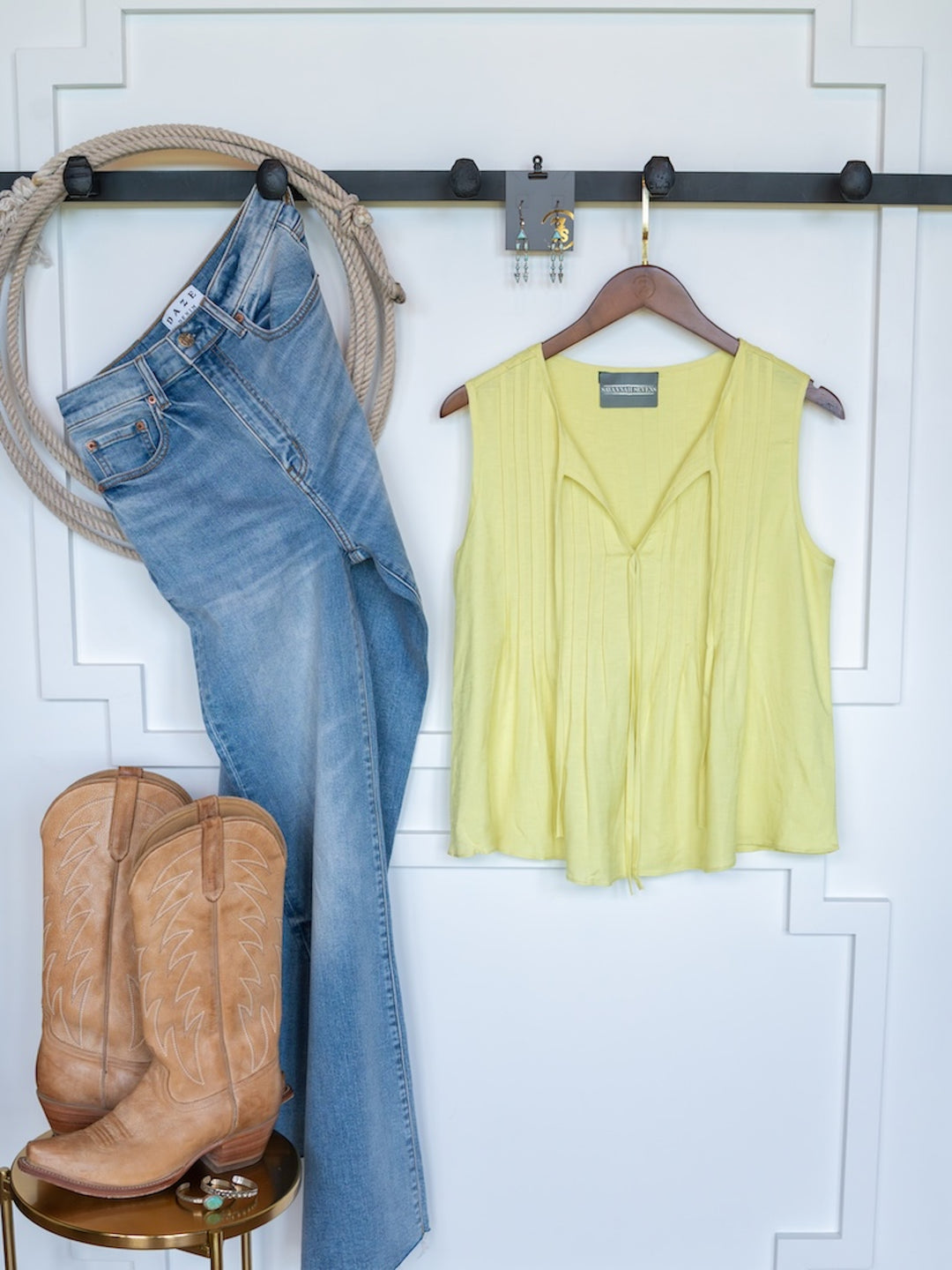 The Marlette by Savannah Sevens western life{&}style: blue jeans, a linen blend sleeveless tie-neck top on a hanger, tan cowboy boots, hoop earrings, and coiled rope displayed on a black rail against a white wall with decorative trim.