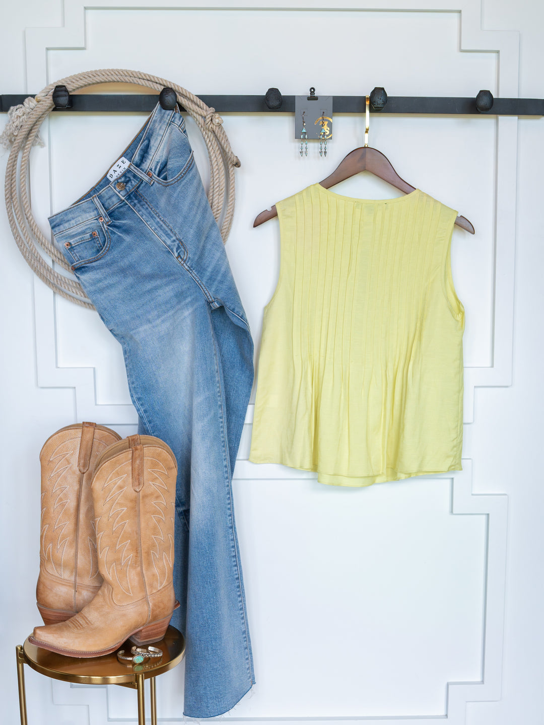The Marlette jeans by Savannah Sevens western life{&}style hang with a linen blend sleeveless top on a wall rack, while tan cowboy boots sit on a round stool, creating a casual western-inspired look.
