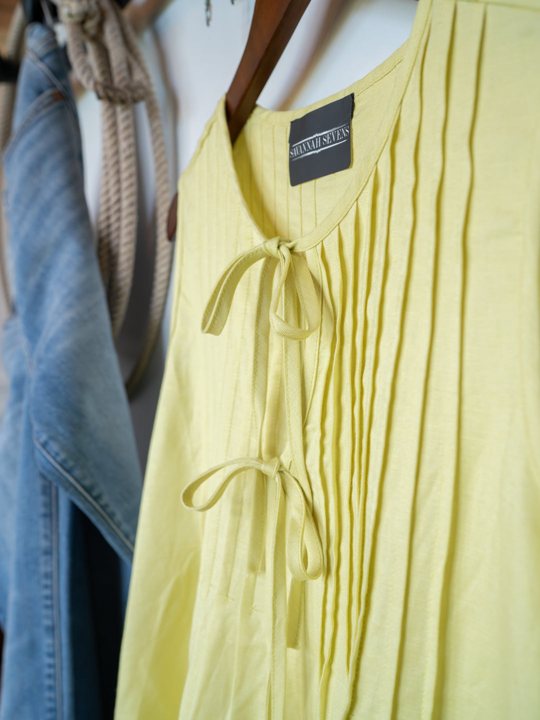 The Marlette pleat top by Savannah Sevens western life{&}style, in chartreuse with an adjustable tie neckline, hangs on a wooden hanger beside light blue denim jeans and a coiled rope.