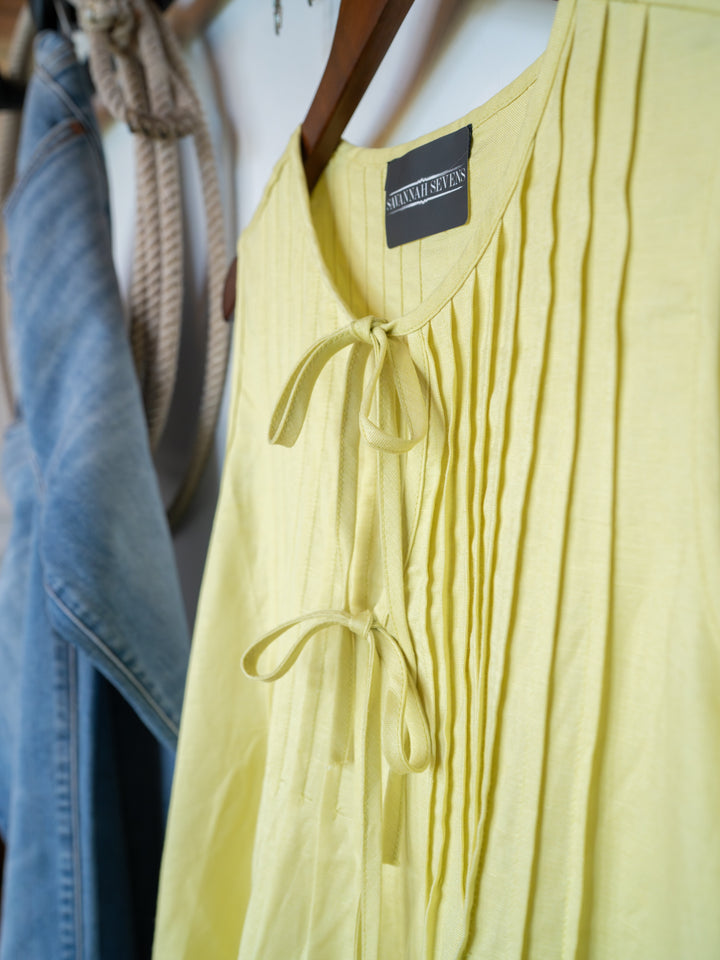 The Marlette pleat top by Savannah Sevens western life{&}style, in chartreuse with an adjustable tie neckline, hangs on a wooden hanger beside light blue denim jeans and a coiled rope.