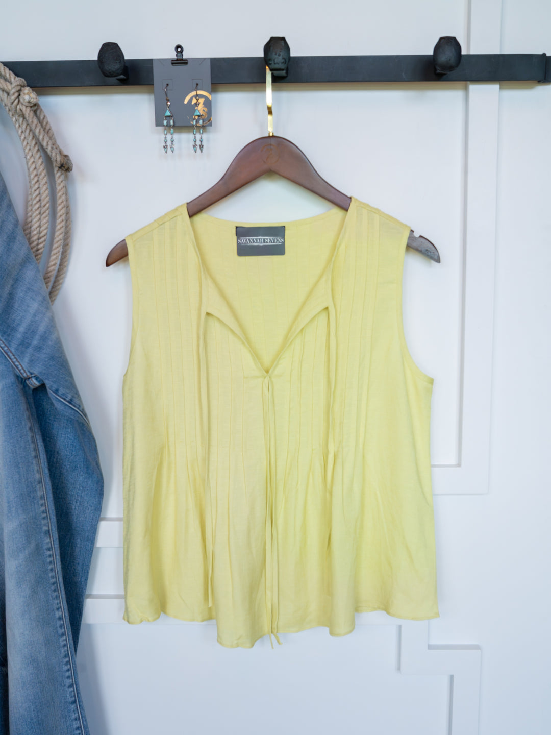 The Marlette by Savannah Sevens western life{&}style, a chartreuse pleat top with an adjustable tie neckline, hangs on a wooden hanger against a white wall. A denim jacket and earrings are also visible.